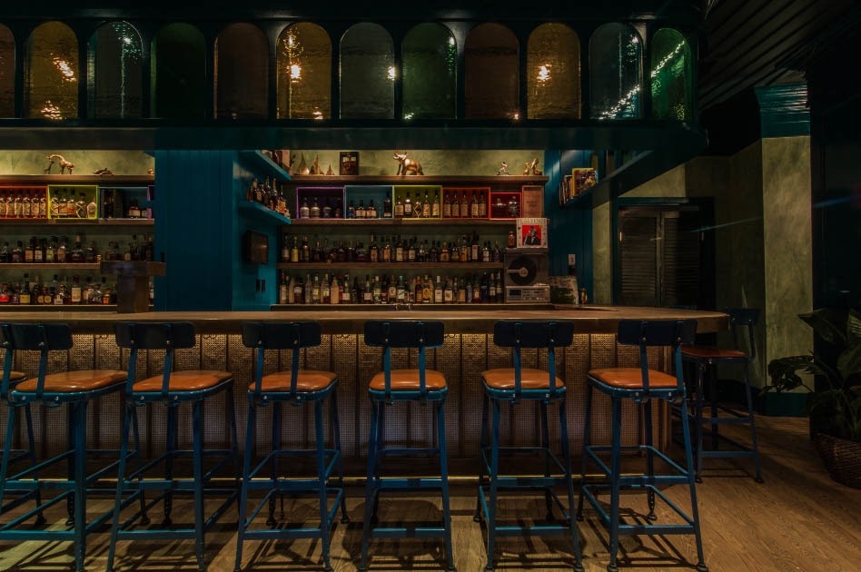 Blue bar stools sit underneath a wooden bar top with a bar with shelves filled with liquor bottles in the back